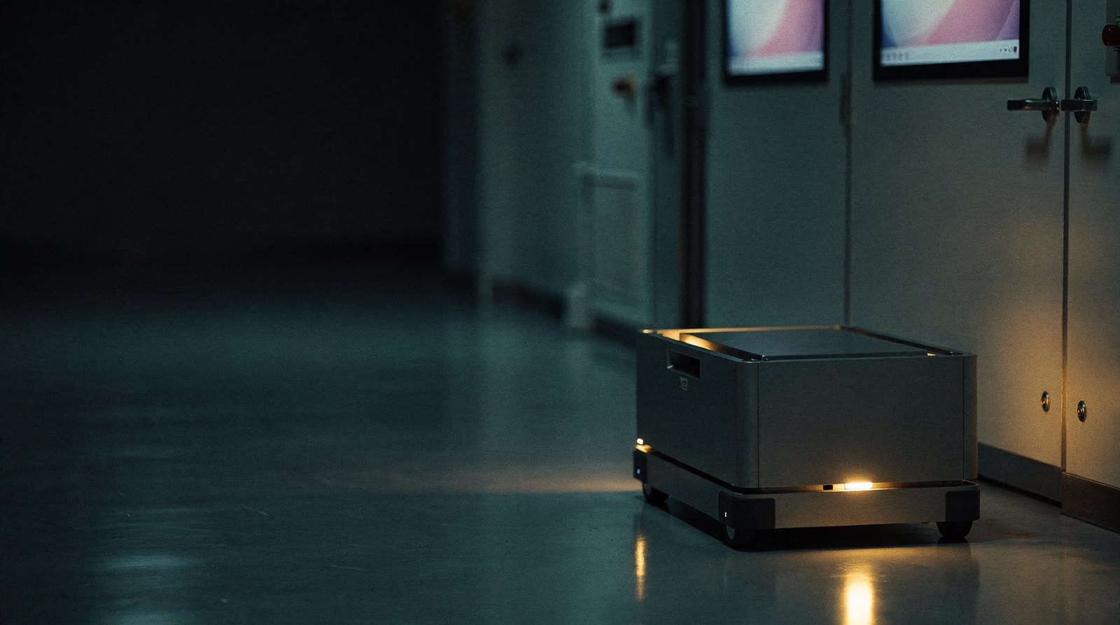 An automated guided vehicle carrying a carrier pod along an empty fab corridor at night, the pod lit warm gold