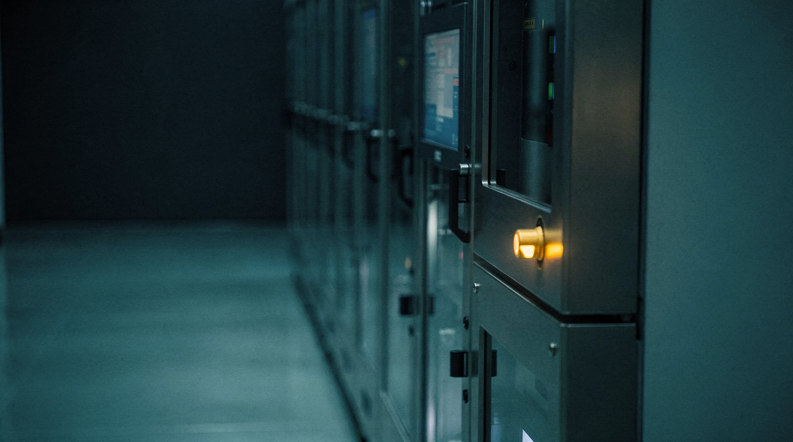 A cleanroom aisle of tool fronts receding into haze, every status lamp dark except one warm gold lamp at the far end