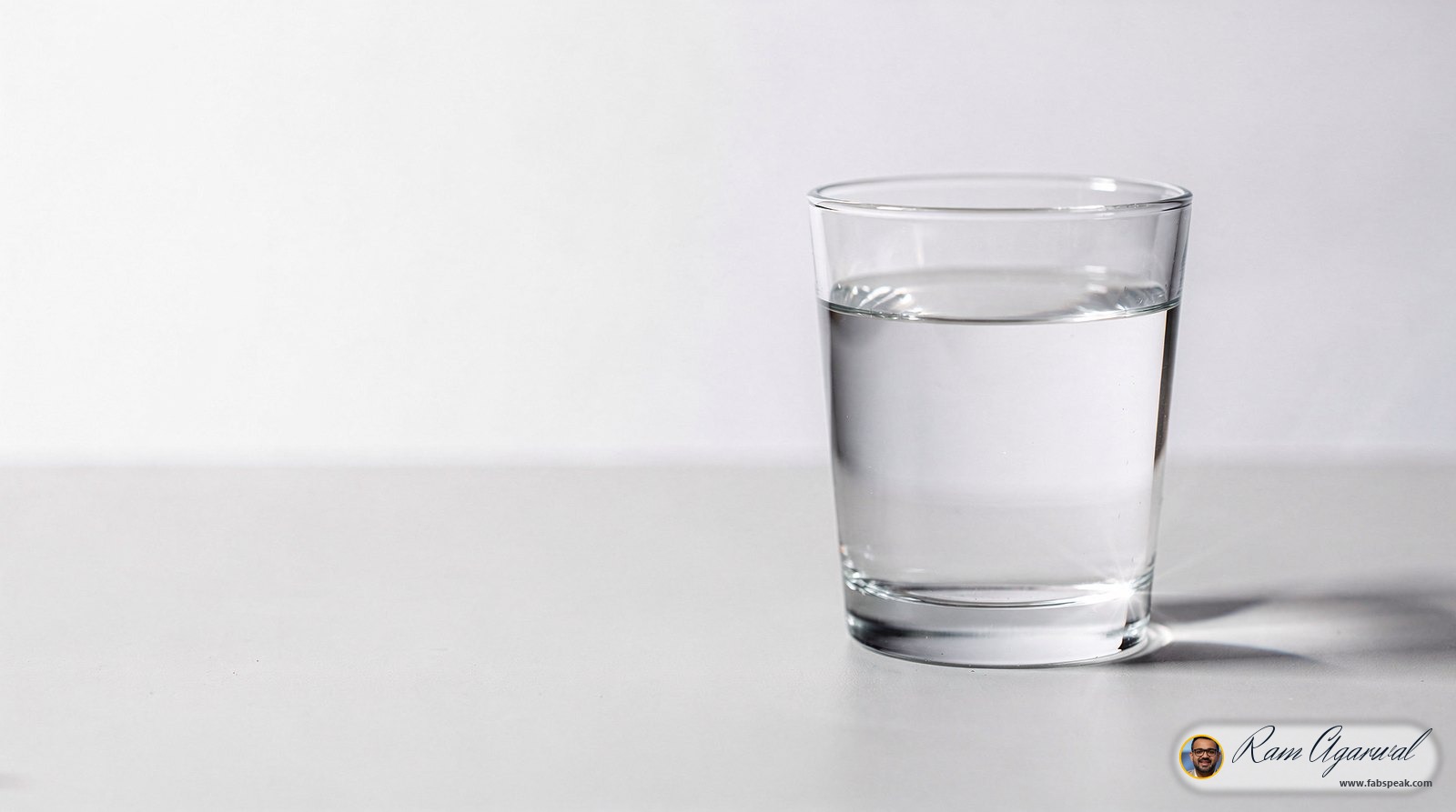 One plain, unlabelled drinking glass filled with perfectly clear water, standing alone on a clean pale grey surface in soft natural daylight. the water is flawlessly transparent and throws a single bright caustic of light on the surface beside the glass. nothing else in the frame at all: no table setting, no jug, no droplets, no background objects. enormous calm empty space around it